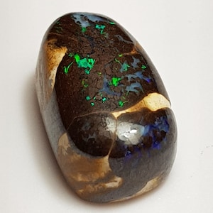 Könnte beinhalten: Ein polierter, oval geformter Opal-Edelstein. Der Stein hat eine dunkelbraune Basis mit lebendigen grünen und blauen Blitzen. Bereiche mit beiger und cremefarbener Matrix sind sichtbar, was zum einzigartigen Aussehen des Steins beiträgt. Der Opal ist vor einem weißen Hintergrund platziert.