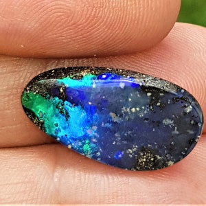 Pode incluir: Opala oval polida, com uma vibrante exibição de cores. A pedra preciosa apresenta uma base escura com flashes de azul, verde e preto. A opala é segurada entre os dedos, mostrando sua beleza natural e padrão único.