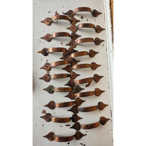 May include: A collection of fourteen copper-toned metal drawer pulls arranged in rows. Each handle features a hammered texture and pointed, spade-shaped mounting plates at both ends, with a single screw hole visible on each mounting plate.