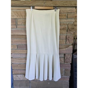 May include: A cream-colored, floor-length skirt featuring a high-waisted design and a flared, ruffled hemline. The skirt is suspended from a wooden hanger, showcasing a smooth, solid-colored fabric with vertical seams and a structured silhouette.