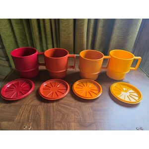 May include: Four stackable mugs with matching lids in a gradient of warm colors, ranging from deep red to bright orange and yellow. Each mug features a square handle and a stepped base design, accompanied by a circular lid with a radial spoke pattern.