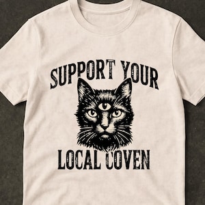 Peut inclure: T-shirt crème avec une illustration noire d'un chat avec un troisième œil. Le texte "SUPPORT YOUR LOCAL COVEN" est imprimé au-dessus et en dessous du graphique du chat en gras noir. Le t-shirt a un col rond et des manches courtes.