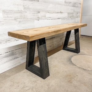 May include: A long wooden bench with a natural finish and black painted A-frame legs. The bench is sitting on a concrete floor.