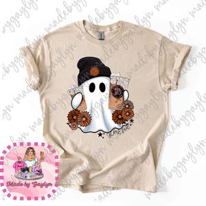 May include: Beige t-shirt featuring a ghost wearing a black beanie and holding two iced coffees. The ghost is surrounded by orange flowers and the text "Ghost Fuel". A black star and text are at the bottom.