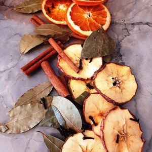 May include: Dried orange slices, apple slices, cinnamon sticks, and bay leaves arranged on a grey marble surface. The ingredients are commonly used in potpourri or for making scented sachets.