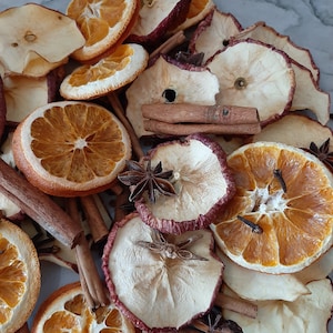 May include: A close-up of a collection of dried fruit and spices, including orange slices, apple rings, cinnamon sticks, and star anise. The ingredients are arranged on a white marble surface.