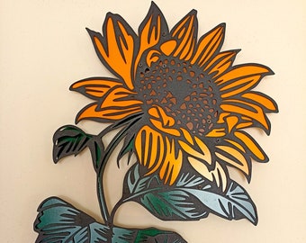 Puede incluir: Un girasol negro y naranja con hojas verdes, recortado en papel y montado sobre un fondo beige claro.