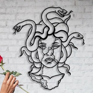 May include: A woman with braided hair is smelling a red rose. Behind her is a black metal wall sculpture of Medusa, a Greek mythological figure with snakes for hair.