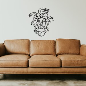May include: A black metal wall sculpture of Medusa, the Greek mythological figure with snakes for hair, is displayed above a brown leather sofa.