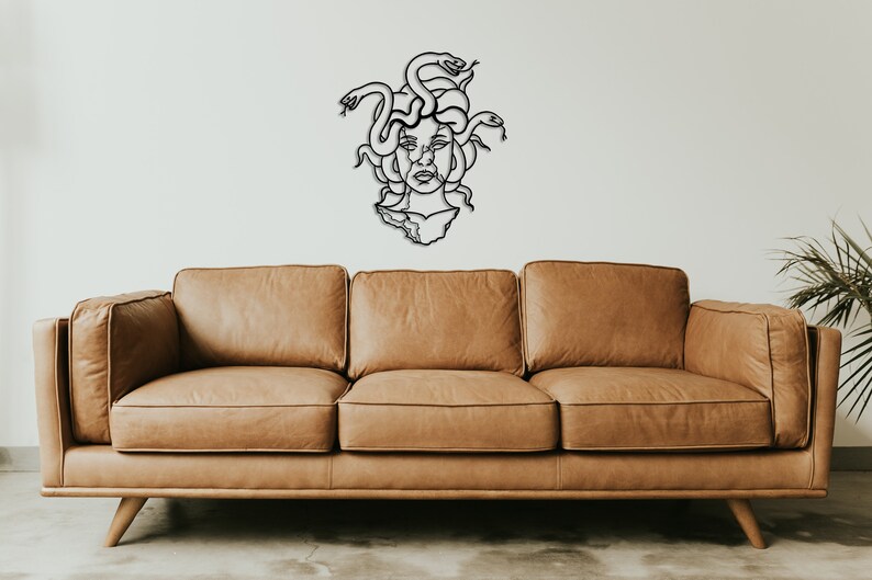 May include: A black metal wall sculpture of Medusa, the Greek mythological figure with snakes for hair, is displayed above a brown leather sofa.