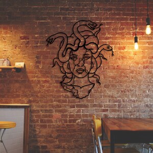 May include: A black metal wall art piece depicting Medusa, a Greek mythological figure with snakes for hair, against a brick wall background.