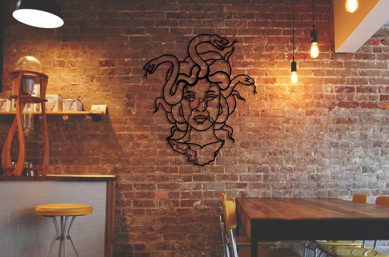 May include: A black metal wall art piece depicting Medusa, a Greek mythological figure with snakes for hair, against a brick wall background.