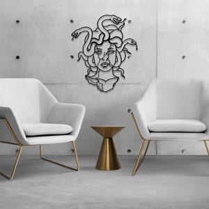 May include: A black metal wall sculpture of Medusa, a mythical Greek figure with snakes for hair, is displayed against a concrete wall. Two white chairs with gold frames and a gold side table are in the foreground.