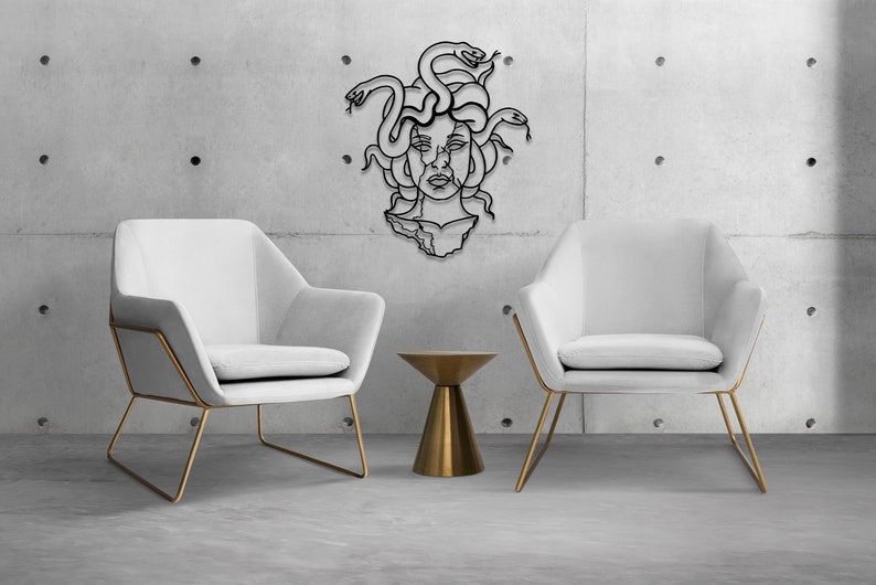 May include: A black metal wall sculpture of Medusa, a mythical Greek figure with snakes for hair, is displayed against a concrete wall. Two white chairs with gold frames and a gold side table are in the foreground.