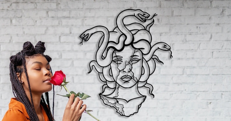 May include: A woman with braided hair is smelling a red rose. Behind her is a black metal wall sculpture of Medusa, a Greek mythological figure with snakes for hair.