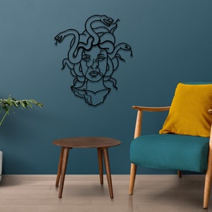 May include: Black metal wall art depicting Medusa, a mythical Greek figure with snakes for hair. The art is mounted on a blue wall, with a teal armchair and a wooden side table in the foreground.