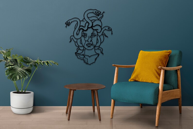 May include: Black metal wall art depicting Medusa, a mythical Greek figure with snakes for hair. The art is mounted on a blue wall, with a teal armchair and a wooden side table in the foreground.