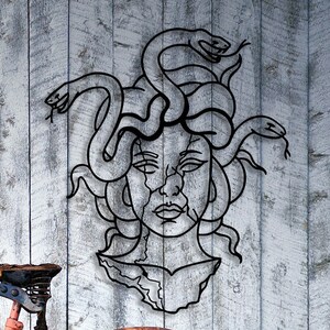 May include: Black metal wall art depicting Medusa, a mythical Greek figure with snakes for hair. The art is in a minimalist style and features a detailed face with a fierce expression.