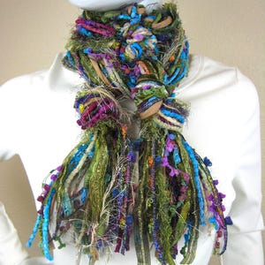 May include: A multi-textured scarf featuring a vibrant mix of olive green, bright blue, magenta, and metallic gold ribbons and yarns. The scarf is constructed from various frayed fabric strips, knotted sections, and looped threads in a dense design.