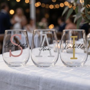 May include: Three clear stemless wine glasses with custom names and initials. The first glass has the name "Scarlett" with a pink "S". The second glass has the name "Ava" with a grey "A". The third glass has the name "Isabella" with a gold "I".