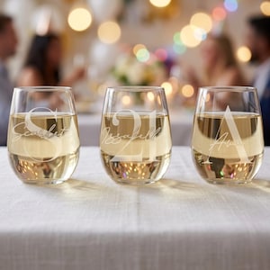 May include: Three clear stemless wine glasses filled with white wine. Each glass features a large white letter and a name in cursive script: S with Scarlett, 21 with Issabell, and A with Ava. The glasses are arranged in a row on a white tablecloth.