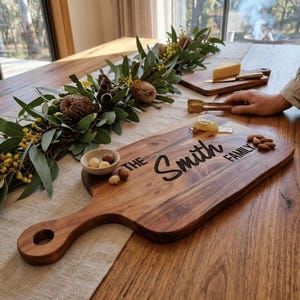 May include: A wooden serving board with a handle, engraved with "The Smith Family" in black script. The board is surrounded by a small bowl of nuts, almonds, and a sprig of greenery. A smaller board with cheese and a knife is in the background.