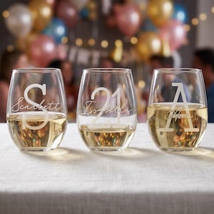 Könnte beinhalten: Drei bauchige Weingläser, gefüllt mit einer hellen Flüssigkeit, wahrscheinlich Wein. Jedes Glas ist mit einem anderen Design graviert: dem Buchstaben "S" und dem Namen "Scarlett", der Zahl "21" und dem Namen "Isabella" sowie dem Buchstaben "A" und dem Namen "Anna".