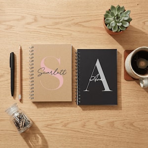 May include: Two spiral-bound notebooks with personalised covers. The left notebook is tan with a large pink letter S and the name Scarlett. The right notebook is black with a large grey letter A and the name Ava. Includes a pen, pencil, and paper clips.