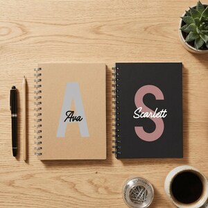 May include: Two spiral-bound notebooks, one tan with a large grey letter A and the name Ava, and one black with a large dusty pink letter S and the name Scarlett. A black pen, a wooden pencil, a small succulent, and a glass jar are present.