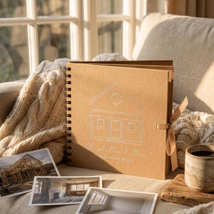 May include: A square, spiral-bound scrapbook with a tan cover featuring a white line drawing of a house, the text Charlotte & Ben, and the words NEW HOME BUILD. A tan ribbon is attached to the side, and three photographs are placed in front.