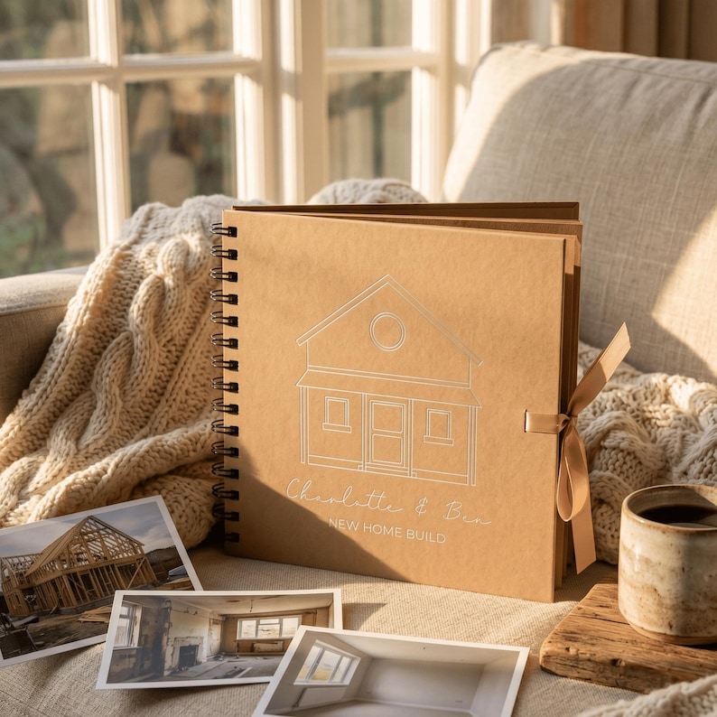 May include: A square, spiral-bound scrapbook with a tan cover featuring a white line drawing of a house, the text Charlotte & Ben, and the words NEW HOME BUILD. A tan ribbon is attached to the side, and three photographs are placed in front.