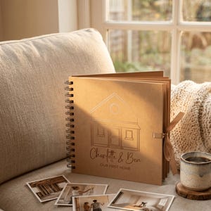 May include: A square, spiral-bound scrapbook with a tan cover featuring a line-art illustration of a house, the names Charlotte & Ben, and the text OUR FIRST HOME. A tan ribbon is tied on the side, and four square photographs lie in front.