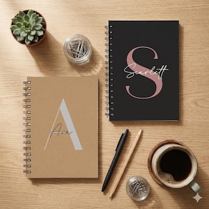 May include: Two spiral-bound notebooks with personalised covers. One is tan with a large grey letter A and the name Ava. The other is black with a large dusty pink letter S and the name Scarlett. Includes a black pen, a wooden pencil, and two small glass jars filled with silver paper clips.