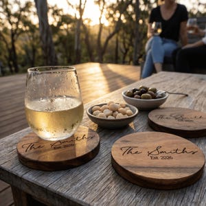 May include: A glass of white wine rests on a wooden coaster engraved with "The Smiths Est 2026". Bowls of olives and nuts are on the wooden table. The scene is set outdoors.