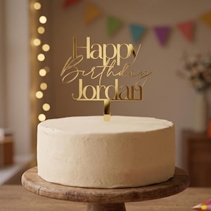 May include: A gold-coloured acrylic cake topper featuring the text Happy Birthday Jordan in a combination of block and script fonts. The topper is positioned vertically on top of a round, smooth, light-coloured cake on a wooden stand.