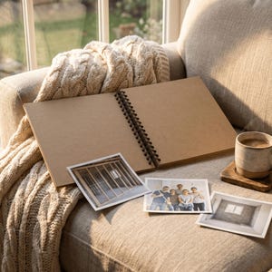 May include: An open spiral-bound sketchbook with blank tan pages, three square photographs featuring a construction frame, a group of five people, and an empty room, and a ceramic mug filled with a brown beverage on a wooden coaster.