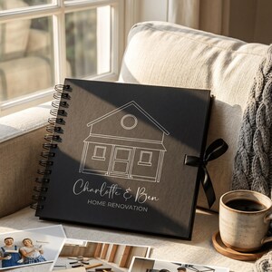 May include: A black square spiral-bound photo album with a white line drawing of a house on the cover. The text reads Charlotte & Ben HOME RENOVATION. A black ribbon tie is attached to the side. The album is placed on a textured fabric surface.