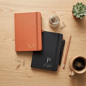 May include: A terracotta orange notebook with the gold initials K.S. and a black notebook with the gold letter P and the name Peyton in script. Both notebooks feature elastic closures and are accompanied by a black pen and a wooden pencil.