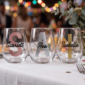 May include: Three clear stemless wine glasses, each with a name: "Scarlett", "Ava", and "Isabella". The letters are pink, silver, and gold. The names are in cursive. The glasses are on a white tablecloth.