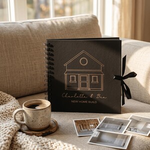 May include: A square black spiral-bound photo album featuring a gold line-art illustration of a house, the names Charlotte & Ben, and the text NEW HOME BUILD. A black ribbon tie is attached to the side, accompanied by a speckled ceramic mug.