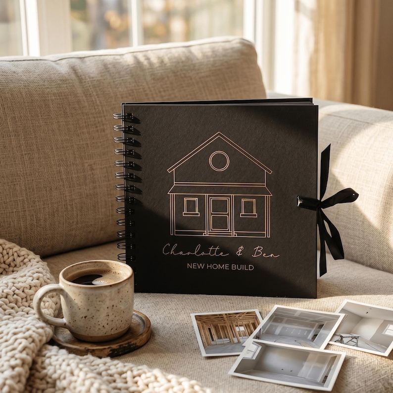 May include: A square black spiral-bound photo album featuring a gold line-art illustration of a house, the names Charlotte & Ben, and the text NEW HOME BUILD. A black ribbon tie is attached to the side, accompanied by a speckled ceramic mug.