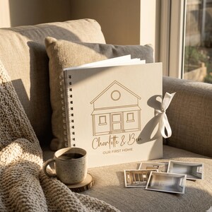 May include: A square, cream-colored spiral-bound scrapbook featuring a gold-toned line drawing of a house, the names "Charlotte & Ben," and the text "OUR FIRST HOME." A white ribbon is tied to the side, and a speckled mug sits nearby.