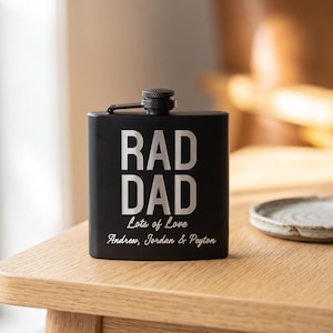 May include: A matte black metal flask featuring the engraved text RAD DAD in a bold sans-serif font, followed by the script text Lots of Love and the names Andrew, Jordan & Peyton. The flask has a hinged screw-top cap at the centre top.