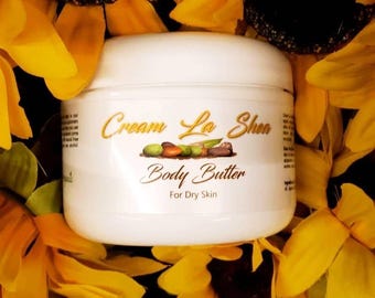 Op de afbeelding: Een witte pot met body butter met het label "Cream La Shea Body Butter For Dry Skin". De pot is omringd door gele zonnebloemachtige bloemen.