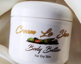 Op de afbeelding: Witte pot met Cream La Shea Body Butter voor droge huid. De pot heeft een gouden etiket met een bruine afbeelding van olijven en amandelen.