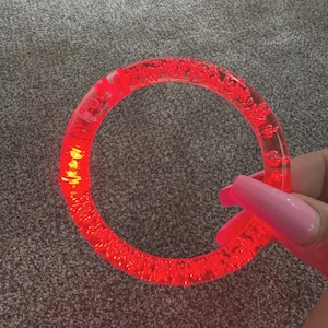 May include: A clear, red, circular bracelet with a textured surface. The bracelet is illuminated with a bright red light. The bracelet is held by a hand with long, pink nails.