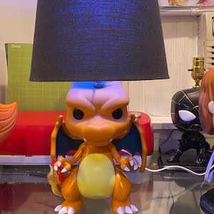 Puede incluir: Una lámpara con temática de Charizard, con una figura naranja y amarilla y una pantalla negra. La figura tiene alas azules y sostiene una llama. La lámpara está sobre una superficie de vidrio.