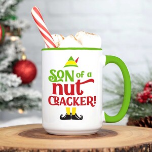 Pode incluir: Caneca de cerâmica branca com borda e alça verdes. A caneca tem um chapéu de duende verde e o texto "SON of a nut CRACKER!" impresso nela. A caneca está cheia de marshmallows e um bastão de caramelo.