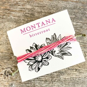 Puede incluir: Una tarjeta blanca con el texto "MONTANA bitterroot" impreso en morado. La tarjeta está atada con una cuerda rosa y blanca y presenta una ilustración en blanco y negro de dos flores.