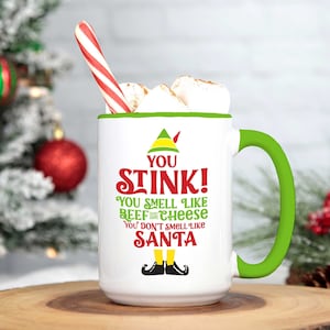 May include: White ceramic coffee mug with a green handle. The mug has a green and red elf design with the text "You Stink! You smell like beef and cheese You don't smell like Santa".